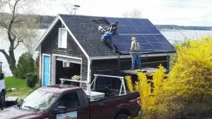 Workers install solar panels on a shingled roof by a serene lake in Maine, with a red truck parked in front and vibrant yellow bushes nearby.