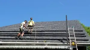 Two construction workers in safety gear stand on a roof under a clear blue sky, handling materials. One worker holds a long beam, preparing for a solar installation in Maine.