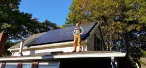 A person stands on a house roof with solar panels installed amidst the lush trees, embracing the eco-friendly innovation of solar in Maine.