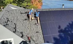 Two workers install solar panels on a sloped roof near a body of water, surrounded by trees.