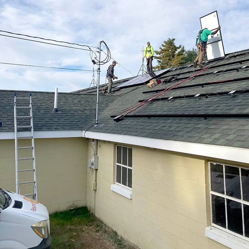 Three workers on a house roof install solar panels, with ladders, cables, and a van visible nearby under a partly cloudy sky.